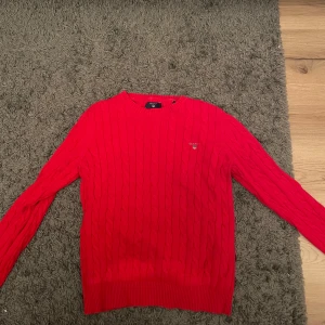 Röd stickad tröja från Gant - Säljer en snygg röd stickad tröja från Gant i premium bomull. Tröjan har ett klassiskt kabelstickat mönster och rund halsringning. Jag har använt den en gång men insåg att den var lite för liten för mig!