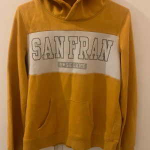 Gul hoodie från H&M med San Fran-tryck - Säljer en gul hoodie från H&M med texten 'San Fran' tryckt på framsidan. Tröjan har en stor ficka framtill och en bekväm huva. Strl 158/164. Perfekt för en avslappnad stil.