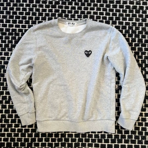 Comme des Garçons play sweatshirt - Säljer en stilren grå sweatshirt från Comme des Garçons med det ikoniska hjärtemärket på bröstet. Tröjan har långa ärmar och ribbade muddar vid ärmslut och nederkant. Perfekt för en avslappnad look. passar xs-s