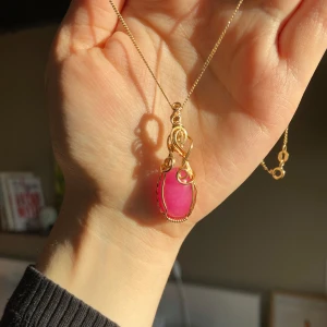 Handgjord berlock med en rosa sten - Handgjord berlock till halsband med en stor rosa sten infattad i dekorativ guldtråd. Hänget har en design med snirkliga detaljer som ger ett konstnärligt intryck. Perfekt för att ge en färgklick till din outfit. Gjord med en koppartråd med guldig anti-tarnish behandling som hindrar tråden från att oxidera och tappa lyster i förtid. (Nickelfri) Bör undvika vatten. 🌻Stenen är en rosafärgad kvarts!