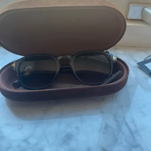 Svarta solglasögon från Tom Ford - Snygga bruna solglasögon från Tom Ford med en elegant design. Glasögonen har en rund form och detaljer i guld på sidorna. Perfekta för att ge en stilren touch till din look. Kommer med ett brunt fodral för säker förvaring.