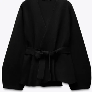 Svart omlottjacka med bälte - Elegant svart omlott tröja med knytbälte i midjan. Tröjan har en stilren design med långa ärmar och en v-ringad öppning. Perfekt för en chic och minimalistisk look.