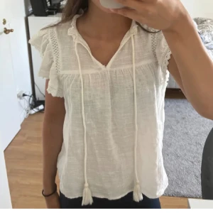 Blus - Säljer en söt vit blus med volanger vid ärmarna. Blusen har korta ärmar och en v-ringad design med knytband framtill. Perfekt för en somrig look!