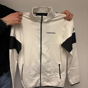 Vit och svart jacka från Peak Performance - Säljer en stilren vit jacka från Peak Performance med svarta detaljer. Jackan har dragkedja framtill och två fickor med dragkedjor. Perfekt för en sportig look.
