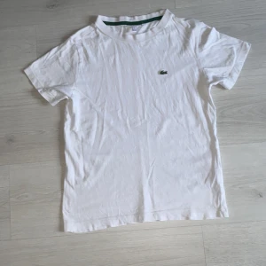 Vit kortärmad t-shirt från Lacoste - Säljer en klassisk vit t-shirt från Lacoste med korta ärmar och det ikoniska gröna krokodilemblem på bröstet. Perfekt för en stilren och avslappnad look. T-shirten har en grön rand på insidan av kragen.
