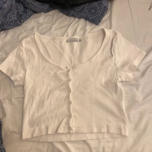 Vit ribbad kortärmad topp - Säljer en vit ribbad topp med korta ärmar och knappar framtill. Toppen är croppad och har en rund halsringning. Perfekt för en avslappnad stil.