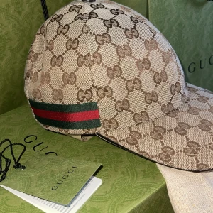 NY keps från Gucci - Snygg beige keps från Gucci med det ikoniska GG-mönstret. Kepsen har en grön och röd randdetalj på sidan. Köpt på GUCCI i Stockholm, helt ny! Nypris i butik 5.650 kr. Digitalt kvitto inlagt på sista bilden! 