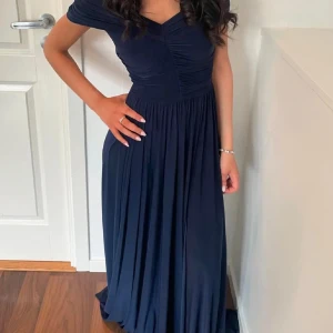 Marinblå långklänning - Elegant marinblå långklänning med offshoulder-design och v-ringning. Klänningen har en smickrande slits framtill som ger en extra touch av stil. Perfekt för speciella tillfällen. Endast använd en gång.