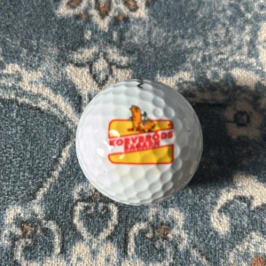 Golfboll från Maxfli med korvbröds bagarn loggan  - Säljer en vit golfboll från Maxfli med en unik korvbröds logga. Bollen har texten 'TOUR FIRE' och siffran 7 tryckt på sig. Perfekt för golfentusiaster som vill ha något annorlunda på banan! 🏌️‍♂️