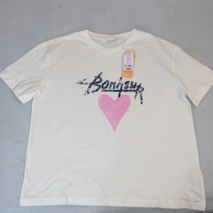 Vit t-shirt med tryck - Säljer en vit t-shirt med ett coolt tryck i form av texten 'Bonjour' och ett rosa hjärta. Perfekt för en avslappnad stil. T-shirten har korta ärmar och en rund halsringning. (Texten och hjärtat ska vara så)