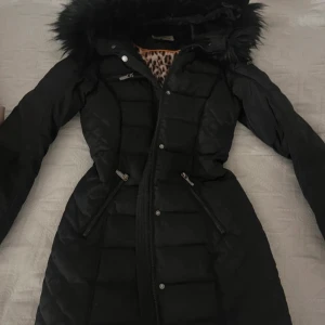 Svart quiltad dunjacka med leopardfoder från Rocco Barocco - Snygg svart dunjacka med quiltat mönster och leopardmönstrat foder. Jackan har en huva med pälsdetaljer och praktiska dragkedjor på fickorna. Perfekt för kyliga dagar. Lite fläckar men syns knappt