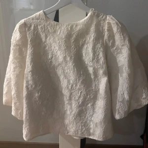 Vit blus från Neo Noir - Säljer en elegant vit blus från Neo Noir med vackert broderat mönster. Blusen har trekvartsärmar och en fin rosettdetalj i ryggen. Perfekt för en stilren look.