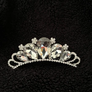Glittrig tiara med stora kristaller - Säljer en elegant tiara med stora, gnistrande kristaller i olika former. Tiaran har en silverfärgad bas och små blommor som dekoration. Perfekt för att ge en kunglig touch till din look. Fästes enkelt med en kam på baksidan.