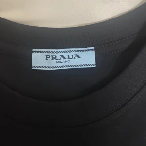 Svart t-shirt från Prada - Snygg svart t-shirt från Prada med en diskret logga i rött på bröstet. Perfekt för en stilren look. T-shirten har en klassisk passform och är kortärmad.