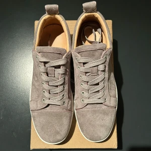 Grå Christian Louboutin skor - Snygga grå sneakers i mocka med den ikoniska röda sulan från Christian Louboutin. Skorna har snörning och en stilren design som passar perfekt för en trendig look. Perfekta för dig som vill ha något unikt och stilrent.