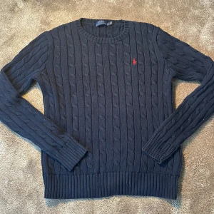 Mörkblå stickad tröja från Polo Ralph Lauren - Säljer en snygg mörkblå stickad tröja från Polo Ralph Lauren med kabelstickat mönster. Tröjan har en broderad röd logga på bröstet och långa ärmar. Perfekt för kyligare dagar!