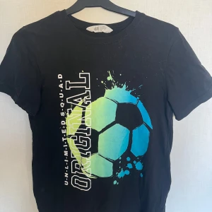 Svart t-shirt med fotbollstryck från H&M - Cool svart t-shirt från H&M med ett stort färgglatt fotbollstryck och texten 'ORIGINAL UNLIMITED SQUAD' på framsidan. Perfekt för fotbollsälskare som vill ha en avslappnad stil.