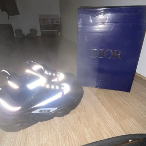 Svarta Dior b22 sneakers med reflekterande detaljer - Chunky svarta Dior sneakers med reflekterande silverdetaljer och meshpaneler. Skorna har kraftig plattformssula, snörning och tydlig Dior-logga på sidan. Kombination av syntet och mesh ger en modern och sportig vibe.