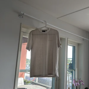 Beige randig t-shirt från Lyle & Scott - Snygg beige t-shirt från Lyle & Scott med diskreta vertikala ränder och klassisk rund hals. På vänster ärm sitter den ikoniska gula fågel-loggan. T-shirten är i mjuk bomull och har en clean, stilren vibe som passar perfekt till jeans eller shorts.