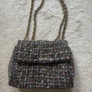 Rutig axelväska från Guess med kedja - Säljer en snygg axelväska från Guess i rutigt, flerfärgat tyg med inslag av svart, vitt, guld, rött och blått. Väskan har en guldfärgad kedjerem och Guess-logga framtill. 