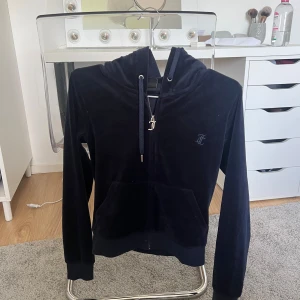 Svart velour hoodie från Juicy Couture - Mysig svart hoodie från Juicy Couture i mjuk velour med dragkedja framtill, huva med snörning och klassisk JC-logga på bröstet. Perfekt för en chill och avslappnad look.