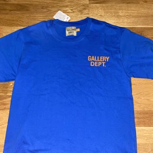 Blå t-shirt Gallery Dept. S, oanvänd. - Blå t-shirt från Gallery Dept. med orange tryck fram och stort tryck på ryggen. Klassisk rund hals och korta ärmar. Skön bomullskvalitet och avslappnad passform. Perfekt statement-plagg för dig som gillar streetwear vibes.
