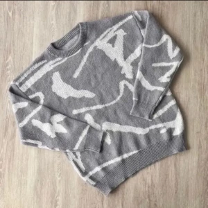 Grå stickad tröja med vitt mönster - Säljer en grå stickad tröja med abstrakt vitt mönster. Tröjan har rund halsringning, ribbade muddar och är långärmad. Perfekt för dig som gillar en chill och trendig look med lite edge.