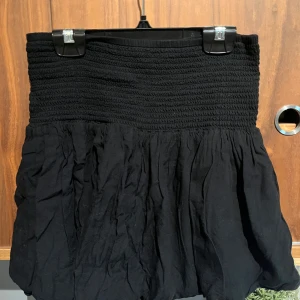 Svart kortkjol med smock - En svart kortkjol med bred smock i midjan och voluminös, rynkad nederdel. Kjolen är enkel men ändå snygg, perfekt för att skapa en chill och trendig look. Tillverkad i mjukt bomullstyg som känns skönt mot huden.