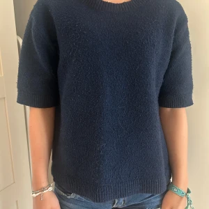 Mörkblå stickad tröja från Kappahl - En enkel och stilren mörkblå stickad tröja från Kappahl. Tröjan har korta ärmar, rund halsringning och ribbade muddar vid ärmslut och nederkant. Perfekt för lager-på-lager och funkar året runt.