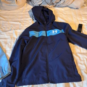Blå vindjacka från Under Armour - Snygg marinblå vindjacka från Under Armour med huva och dragkedja. Jackan har en ljusblå detalj över bröstet och logga på vänster sida. Tillverkad i lätt polyester med meshfoder för extra komfort. Perfekt för dig som gillar sportig stil.