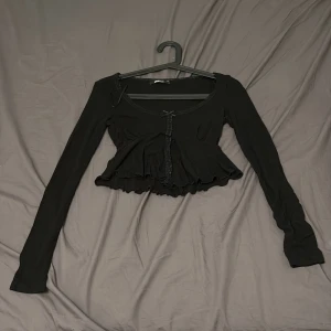 Svart croppad topp från Gina Tricot - Supersöt svart croppad topp från Gina Tricot med långa ärmar, volangkant nertill och spetsdetaljer längs urringningen. Söt liten rosett framtill och figurnära passform. Perfekt för dig som gillar en feminin och trendig look.