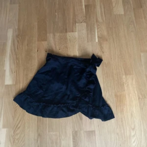 Svart omlottkjol med volang - Supersöt svart kort omlottkjol med volangkant och knyt i sidan. Kjolen har en feminin och trendig look som passar perfekt till sneakers eller boots. Materialet känns mjukt och följsamt, vilket gör den bekväm att bära hela dagen.