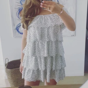 Vit prickig volangklänning offshoulder - Supersöt kort volangklänning i vitt med svarta prickar. Offshoulder-modell med flera lager volanger som ger en fluffig och lekfull look. Perfekt för sommarens varma dagar och har en luftig känsla. Klänningen är ärmlös och har en avslappnad passform.