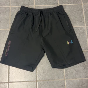 Svarta träningsshorts från Under Armour - Svarta träningsshorts från Under Armour med regnbågsfärgad logga och text på ena benet. Shortsen har elastisk midja med snörning och två sidofickor. Lätt och luftig modell i syntetmaterial, perfekt för gym eller löpning.