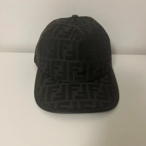 Svart Fendi keps med FF-mönster - Snygg svart keps från Fendi med ikoniskt FF-monogram över hela kepsen. Klassisk form med böjd skärm och justerbar rem bak där Fendi-loggan syns. Tillverkad i ett slitstarkt tyg med exklusiv känsla, perfekt för dig som gillar designeraccessoarer.