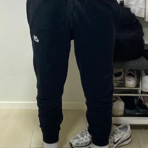 Svarta mjukisbyxor från Nike - Svarta mjukisbyxor från Nike med broderad vit logga på vänster lår. Byxorna har dragsko i midjan och muddar vid bensluten för en skön och avslappnad passform. Perfekta för chill eller träning. Tillverkade i mjukt bomullsmaterial.