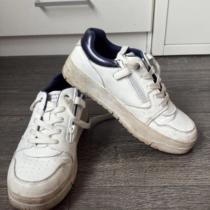 Vita sneakers från Tommy Hilfiger - Snygga vita sneakers från Tommy Hilfiger med marinblå detaljer vid hälen. Skorna har snörning, dragkedja på sidan och en platt sula. Perfekta för dig som gillar en clean och sportig stil. Tillverkade i skinn och syntet.