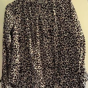 Leopardmönstrad skjorta från Lindex - Snygg långärmad skjorta från Lindex med leopardmönster i beige och svart. Skjortan har klassisk krage, knappar framtill och är tillverkad i ett mjukt, lätt material som känns skönt mot huden. Perfekt för dig som vill sticka ut med en trendig look.