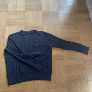 Mörkblå kabelstickad tröja från Gant - Snygg mörkblå kabelstickad tröja från Gant med rund hals och diskret logga på bröstet. Tröjan har långa ärmar och ribbade muddar vid ärmslut och nederkant. Perfekt för dig som gillar klassisk och stilren look.