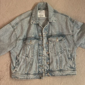 Ljusblå jeansjacka från Pull&Bear - Säljer en klassisk ljusblå jeansjacka från Pull&Bear. Jackan har en oversized passform, knappar framtill och två bröstfickor med lock. Perfekt att slänga över en hoodie eller t-shirt för en chill look.