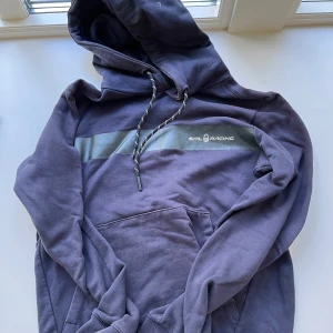 Mörkblå hoodie från Sail Racing - Säljer en mörkblå hoodie från Sail Racing med svart logga och bred rand över bröstet. Tröjan har huva med snörning, känguruficka och är tillverkad i mjuk bomull. Perfekt för dig som gillar en clean och sportig stil.