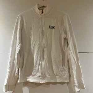 Vit zipjacka från EA7 Emporio Armani - Vit jacka från EA7 Emporio Armani med hel dragkedja och ståkrage. Jackan har två fickor framtill och diskret logga på bröstet. Perfekt för en clean och sportig look. Långärmad och i mjukt bomullsmaterial.