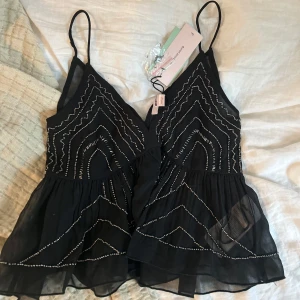 Svart tröja från Gina Tricot Young - Supersnygg svart tröja från Gina Tricot Young med tunna axelband och glittriga silverpärlor i geometriskt mönster. Kjolen är lätt och luftig med volang. Perfekt för dig som vill sticka ut med en cool och trendig look.