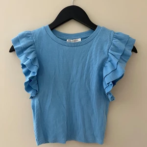 Blå volangtopp från Zara - Supersöt blå topp från Zara med ribbad struktur och volangdetaljer på axlarna. Ärmlös modell som ger en fräsch och trendig look. Perfekt att styla med jeans eller kjol för en cool vibe.
