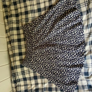 Blommig blå kjol från Lager 157 M/L - Supersöt blå kortkjol från Lindex med vitt blommönster och smock i midjan. Kjolen har en lätt utställd form och är perfekt för dig som gillar en feminin och avslappnad stil. Passar dig som vill ha något bekvämt och snyggt till sommaren.