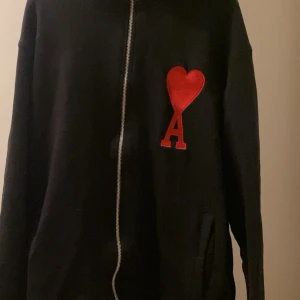 Svart zipjacka från Ami Paris - Svart jacka från Ami Paris i 100% bomull med dragkedja framtill. På bröstet sitter ett stort rött broderat hjärta med bokstaven A. Jackan har fickor på sidorna och en clean, stilren look som passar perfekt till streetwear.Storlek S 