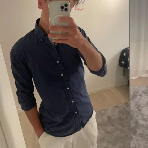 Ralph Lauren skjorta - Säljer en mörkblå skjorta från Ralph Lauren i slim fit-modell. Skjortan är tillverkad i bomullspoplin och har klassisk krage, knäppning framtill och det ikoniska röda Polo-logot broderat på bröstet. Perfekt för en clean och stilren look.