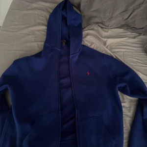 Blå hoodie från Polo Ralph Lauren - Mörkblå hoodie från Polo Ralph Lauren med dragkedja och huva. Klassisk röd logga broderad på bröstet och två fickor framtill. Perfekt för chill dagar eller när du vill ha en clean och enkel stil.