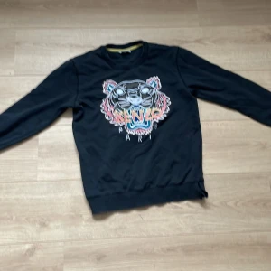 Svart Kenzo sweatshirt med tiger - Svart sweatshirt från Kenzo med ikoniskt broderat tigermotiv i flera färger på bröstet. Tröjan har rund hals, långa ärmar och ribbade muddar. Snygg detalj med liten slits i nederkanten. Perfekt statement piece för dig som gillar streetstyle. Har ett litet hål på sig, kom privat för diskussion av pris😊