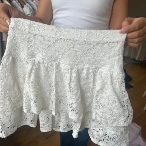 Vit spetskjol med volang - Supersöt vit kortkjol i spets med volangdetaljer. Kjolen har en bred midja och ett romantiskt, feminint uttryck. Perfekt för dig som gillar en ljus och fräsch stil med extra detaljer.